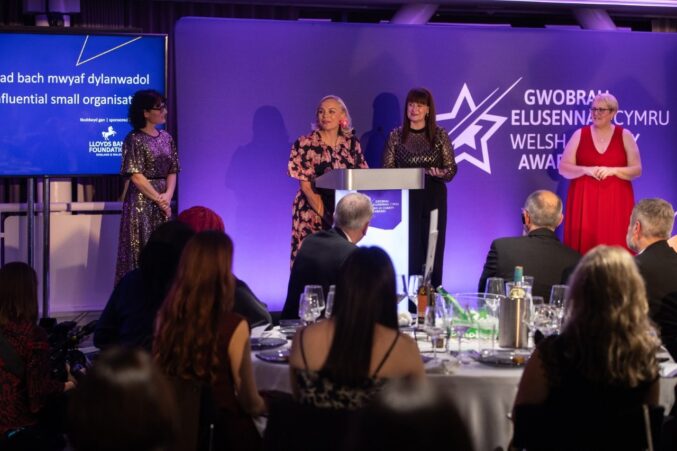 Welsh Charity Awards ceremony with several people standing on stage near a podium. A large purple backdrop displays the event logo and text in Welsh and English. A screen to the left shows the award category for the most influential small organisation. Guests are seated at round tables in the foreground.