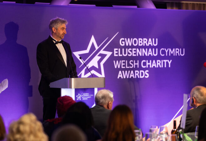 A person speaking at a podium on stage at the Welsh Charity Awards, with the event logo and bilingual text “Gwobrau Elusennau Cymru / Welsh Charity Awards” displayed on a purple backdrop.