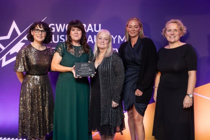 A group of five people standing on a stage at the Welsh Charity Awards. One person in the center is holding an award plaque.