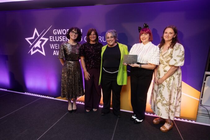A group of five people standing on a stage at the Welsh Charity Awards. One person in the center is holding an award plaque.